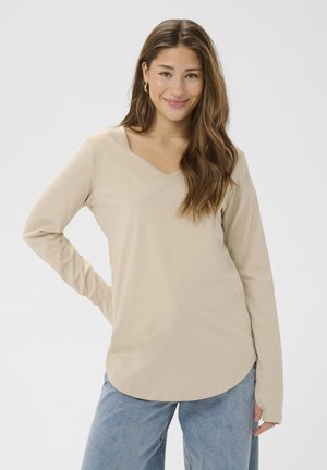 Mladá žena s dlhými hnedými vlasmi, oblečená v béžovom tričku s dlhým rukávom a modrých džínsoch, usmievajúca sa s jednou rukou na bokoch.