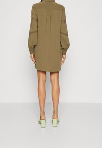 Olivengrünes Hemdkleid mit Puffärmeln, strukturiertem Stoff und einer Rückenschleife. Kombiniert mit mintgrünen Blockabsatzsandalen.