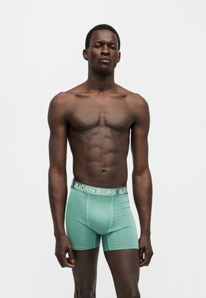 Μυώδης άνδρας στέκεται μπροστά σε απλό φόντο, φορώντας πράσινα μπόξερ Björn Borg, με χαλαρά χέρια στα πλευρά, κοιτάζοντας προς τα εμπρός.