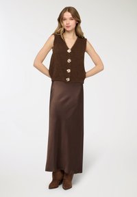 Gilet senza maniche marrone lavorato a maglia con bottoni dorati, abbinato a una lunga gonna di raso marrone e stivali in suede marroni, creando un look a strati.