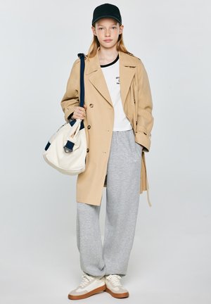 Młoda osoba ubrana w beżowy prochowiec, szare spodnie dresowe, białe trampki, czapkę z daszkiem w czarnym kolorze i niosąca białą torbę typu tote z granatowymi paskami.