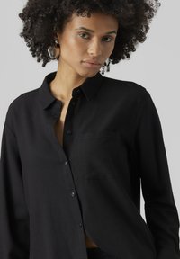 Camicia nera con bottoni, colletto a punta, maniche lunghe e taschino sul petto. Tessuto morbido e liscio con una leggera lucentezza.