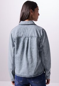 Giacca di jeans blu chiaro con colletto classico, maniche lunghe e spalle rinforzate. Presenta una finitura morbida e leggermente testurizzata e polsini con bottoni.