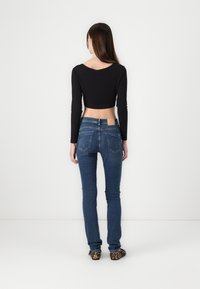 Black cropped long-sleeve top with a low scoop back, paired with fitted blue denim jeans featuring back pockets and a tan label on the waistband.