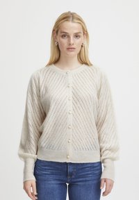 Cardigan en tricot beige avec un motif de rayures diagonales, fermeture à boutons avec des accents en perles, et poignets côtelés, assorti avec un jean bleu.