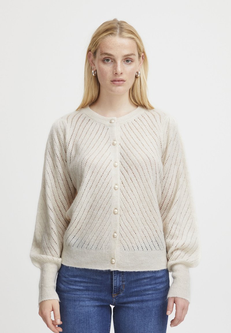 Cardigan en tricot beige avec un motif de rayures diagonales, fermeture à boutons avec des accents en perles, et poignets côtelés, assorti avec un jean bleu.