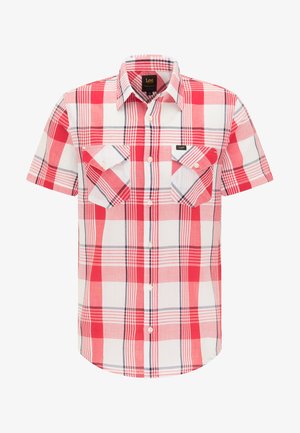 Man draagt een rood-wit geruite korte mouwen overhemd, blauwe jeans en zwarte lage sneakers, staat met gekruiste armen.