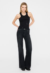 Top senza maniche nero con decorazioni, abbinato a jeans in denim scuro a gamba larga. L'outfit presenta una cintura con una fibbia di metallo argentato.