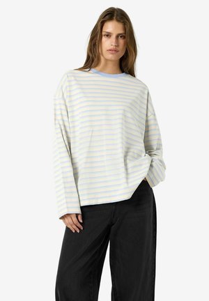Jeune femme portant une chemise jaune clair à manches longues avec de fines rayures horizontales bleues et un pantalon noir ample sur fond blanc.