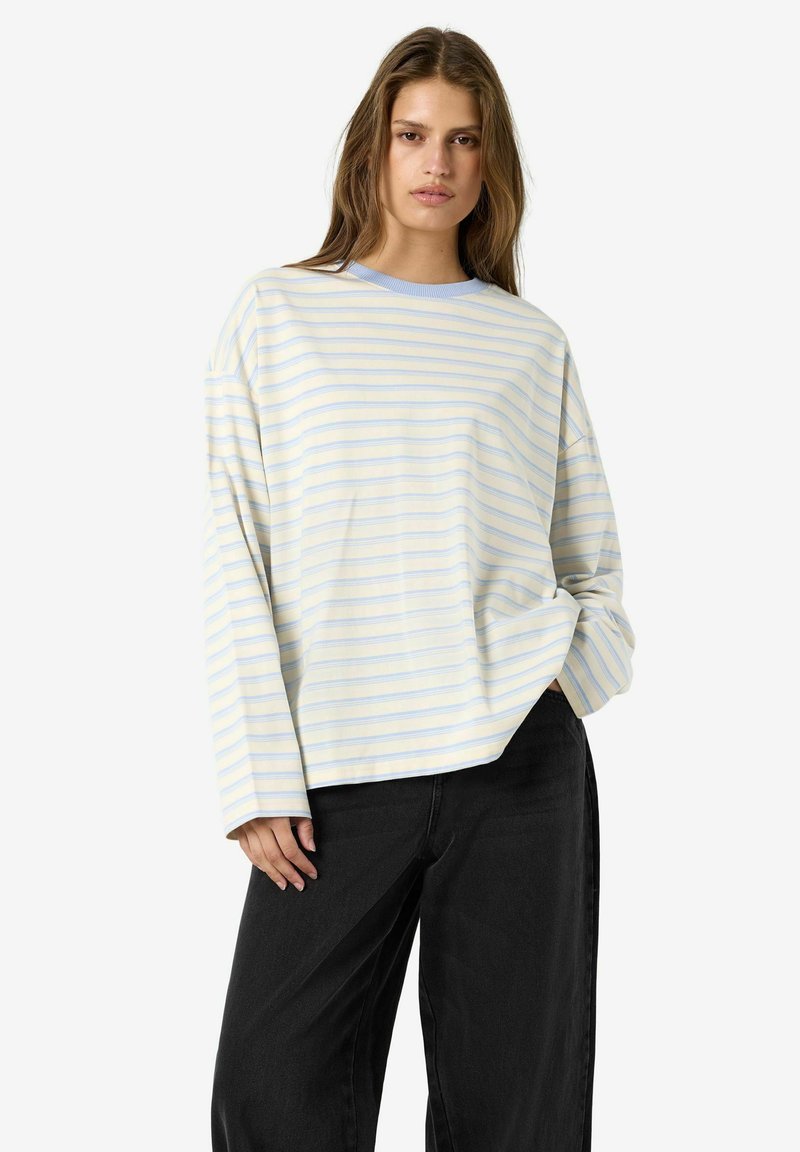 Jeune femme portant une chemise jaune clair à manches longues avec de fines rayures horizontales bleues et un pantalon noir ample sur fond blanc.