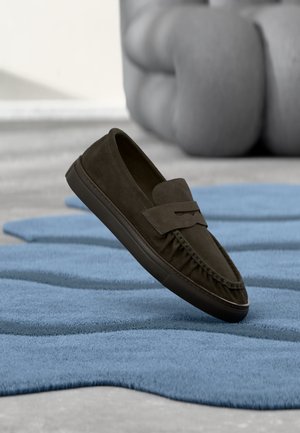 Zapato mocasín de ante marrón oscuro flotando sobre una alfombra azul texturizada con muebles grises esculpidos desenfocados al fondo.