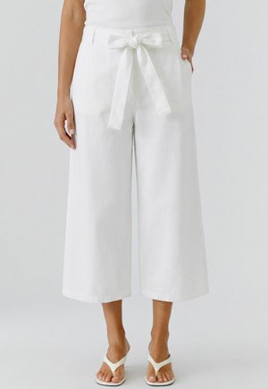 Vrouw draagt witte, wijde cropped broek met hoge taille, een voorgebonden ceintuur en zijzakken, gecombineerd met witte sandalen met hak.