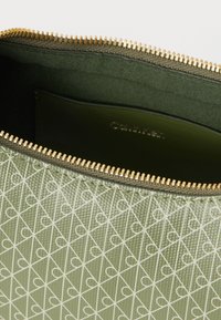 Πράσινη υφασμάτινη θήκη με λευκό γεωμετρικό σχέδιο, χρυστό μεταλλικό φερμουάρ και εσωτερικό ελιάς με ετικέτα μάρκας.