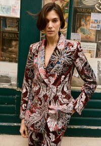 Blazer doppiopetto in velluto con intricati motivi floreali in bordeaux e argento. Presenta audaci accenti blu e rosa, rever a lancia, design aderente.