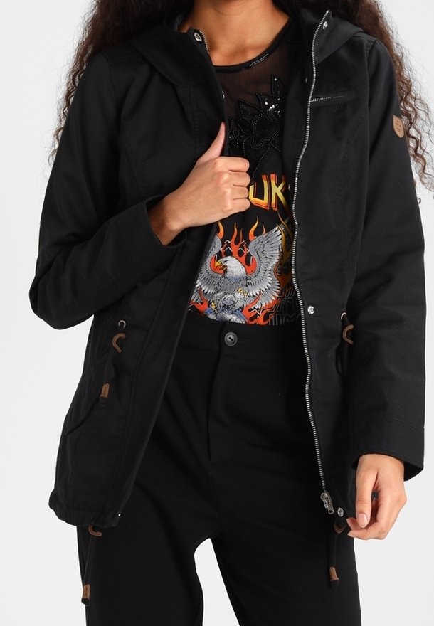 Personne portant une veste noire par-dessus un t-shirt graphique avec un design d'aigle en flammes, assorti à un pantalon noir taille haute.