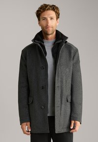 Schwarzgrauer Wollmischmantel mit Fischgrätmuster, Knopfleiste, zwei Fronttaschen und Stehkragen. Innenshirt sichtbar.