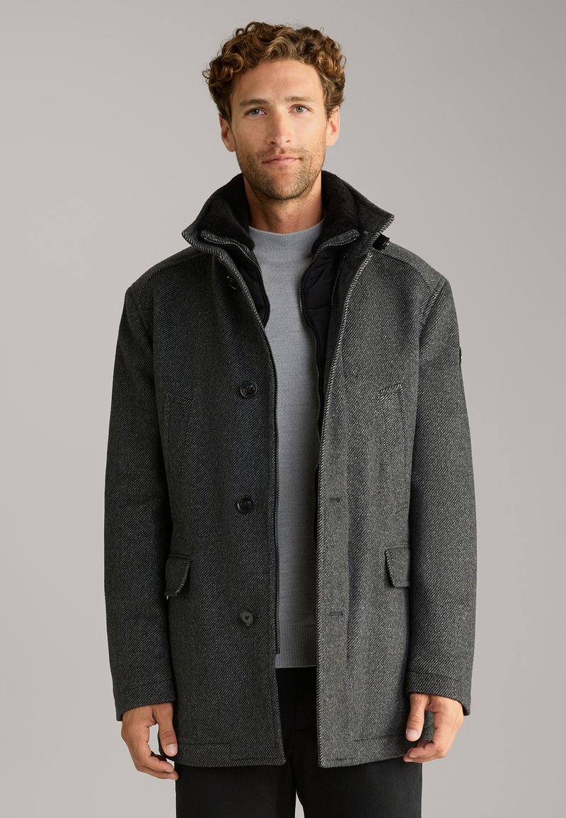 Schwarzgrauer Wollmischmantel mit Fischgrätmuster, Knopfleiste, zwei Fronttaschen und Stehkragen. Innenshirt sichtbar.