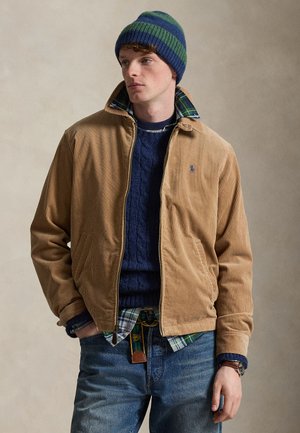 Junger Mann trägt eine beige Cordjacke über einem marineblauen Zopfmuster-Pullover und einem karierten Hemd, mit einer blau-grün gestreiften Mütze und blauen Jeans.