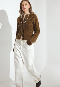 Olīvu zaļa adīta džempera jakas ar apaļu kaklu, pogu aizdari un ribotu apakšmalu, pārī ar baltām platām biksēm un melniem augstiem zābakiem.