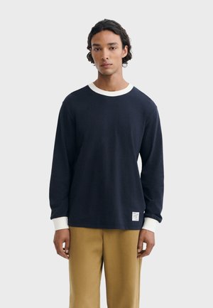 Jeune homme aux cheveux bouclés foncés portant une chemise bleu marine à manches longues avec col et poignets blancs, et un pantalon marron clair, debout devant un fond blanc.
