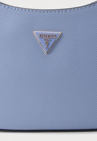 Geantă de mână texturată de culoare albastru deschis, cu un logo triangular auriu și albastru "GUESS USA" în partea din față, având o margine superioară netedă și accente negre.