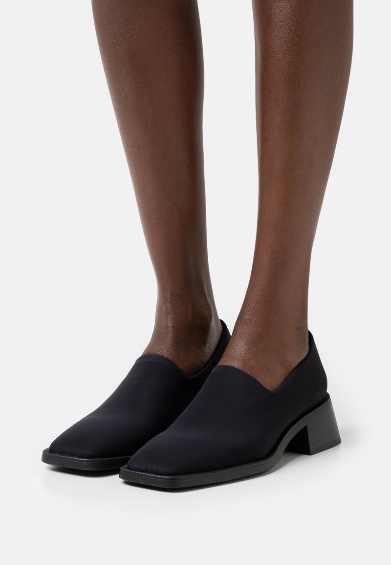 Bottines noires en tissu avec un bout pointu, un petit talon carré et une texture lisse, présentant un design minimaliste.