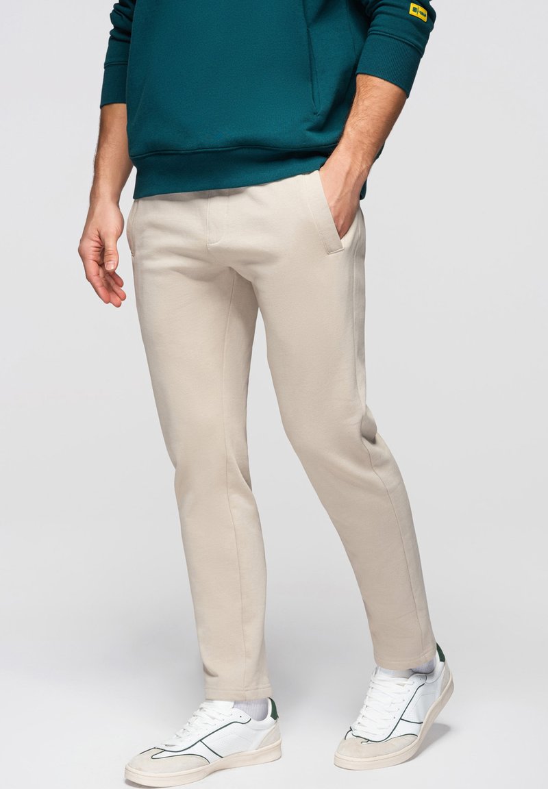 Hombre con pantalones ajustados beige, zapatillas blancas y sudadera azul verdosa, con la mano en el bolsillo, de pie sobre un fondo claro y liso.