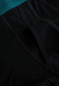 Fekete szövetből készült sportviselet, látható varrásokkal és türkiz texturált derékpánttal.