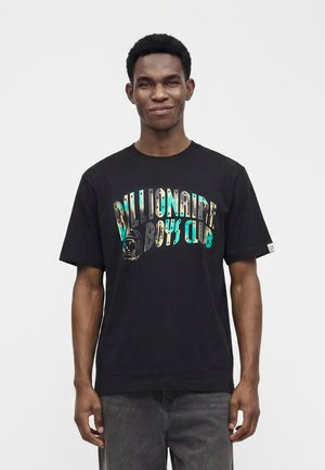 Jeune homme portant un t-shirt noir Billionaire Boys Club avec un texte coloré, debout devant un fond blanc uni.