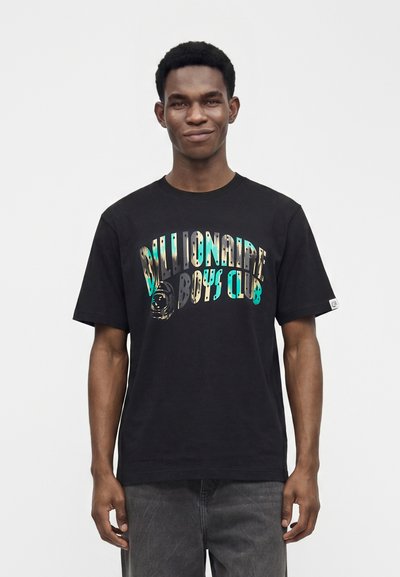 Νέος άνδρας με μαύρο T-shirt Billionaire Boys Club με πολύχρωμο κείμενο, στέκεται μπροστά σε ένα απλό λευκό φόντο.
