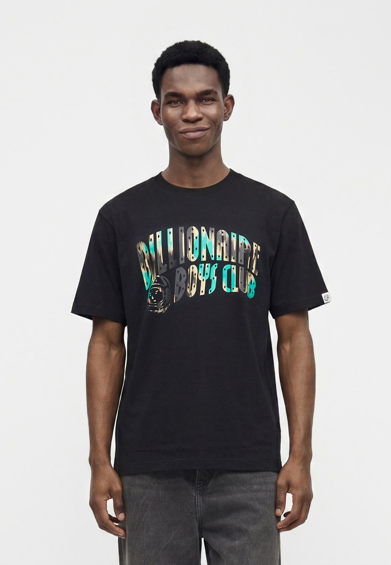 Νέος άνδρας με μαύρο T-shirt Billionaire Boys Club με πολύχρωμο κείμενο, στέκεται μπροστά σε ένα απλό λευκό φόντο.