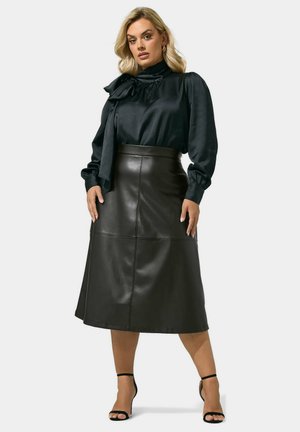 Mujer que lleva una blusa de satén negra con detalle de lazo y una falda midi de cuero negra, combinadas con tacones negros con pulsera en el tobillo, de pie con confianza.