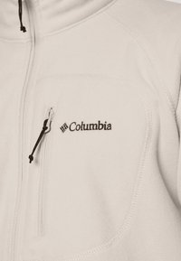 Lys beige fleece jakke med lynlåslomme på brystet og sort Columbia-logo; glat tekstur med flade sømme og justerbar snor.