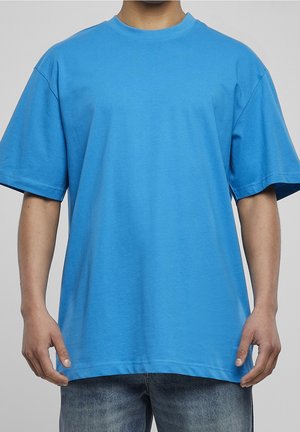 Personne porte un T-shirt ample bleu vif à manches courtes et un jean bleu délavé, debout devant un fond gris uni.