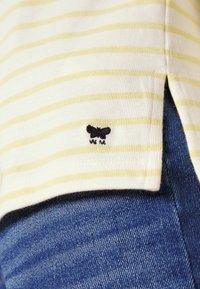 Gestreiftes Baumwoll-T-Shirt in Weiß mit gelben Linien, das eine kleine schwarze Schmetterlingsstickerei nahe dem Saum aufweist, zusammen mit blauem Denimstoff.