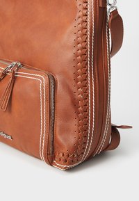 Un sac en cuir marron avec des détails tressés, des coutures blanches contrastantes et une poche zippée à l'avant avec une frange en cuir. Texture lisse.