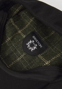 Black cap with a green plaid wool lining. Features a black tag with "Brixton" logo and a crest. Textured fabric with detailed stitching.