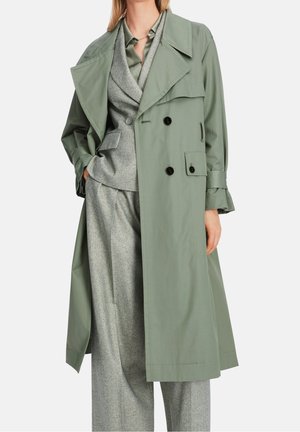 Vrouw die een lange saliegroene trenchcoat draagt over een grijs pak met brede broekspijpen en een blazer, staand met één hand in de zak.