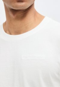 Homme portant un t-shirt blanc à col rond avec un texte discret en relief "Peak Performance" sur la poitrine, sur un fond uni.