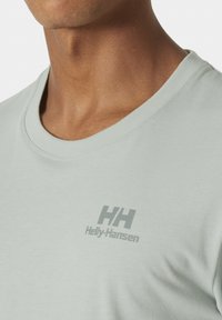 Camiseta de algodón verde claro con cuello redondo y mangas cortas, con un sutil estampado gris del logo en el lado izquierdo del pecho. Textura suave.