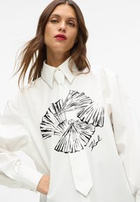 Biała koszula z czarnym wzorem w kształcie wachlarza na środku, z kołnierzykiem na guziki i detalem nawiązującym do krawata z przodu. Gładka tekstura materiału.