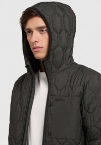 Quilted dark green jacket with a hood, featuring a zippered chest pocket and hexagonal pattern across the fabric, soft texture.