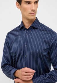 Camicia blu navy con bottoni, caratterizzata da sottili righe verticali bianche, con colletto a punta e maniche lunghe con polsini abbottonati.