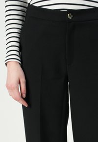 Pantalones negros ajustados con pliegues frontales y un cierre de un solo botón, combinados con una blusa de manga larga a rayas en blanco y negro.