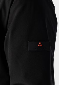 Pantaloni di tessuto nero caratterizzati da una texture liscia, con una toppa nera rettangolare e un logo con tre punti rossi sul lato.