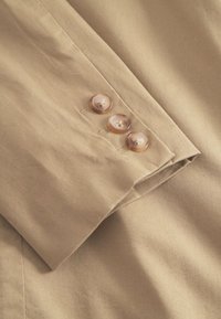 Bež manžeta košulje s tri prozirna gumba, glatke teksture, uredno presavijenim rubom i suptilnim sjajem.