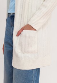Ribbed white cardigan with long sleeves, featuring a front pocket and a soft texture. Worn over blue high-waisted jeans.