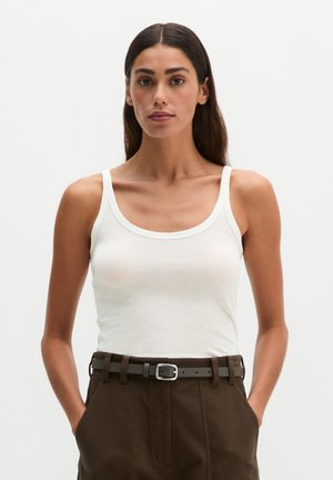 Donna con lunghi capelli scuri che indossa una canottiera bianca infilata in pantaloni marroni a vita alta con una cintura scura, in piedi con le mani in tasca.