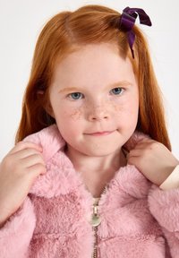 Giacca di pelliccia sintetica rosa chiaro con chiusura a zip e un tirante a forma di cuore, indossata da un bambino con capelli rossi e lentiggini.