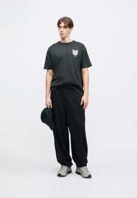 Μαύρο βαμβακερό t-shirt με γραφικό σχέδιο στο στήθος, σε συνδυασμό με χαλαρές μαύρες παντελόνες και γκρι αθλητικά παπούτσια. Ένα μαύρο καπέλο κρατιέται στο αριστερό χέρι.